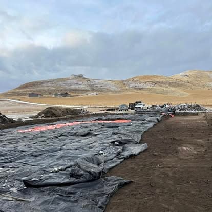 Construction site with tarps spread over earth, surrounded by hills under a cloudy sky.