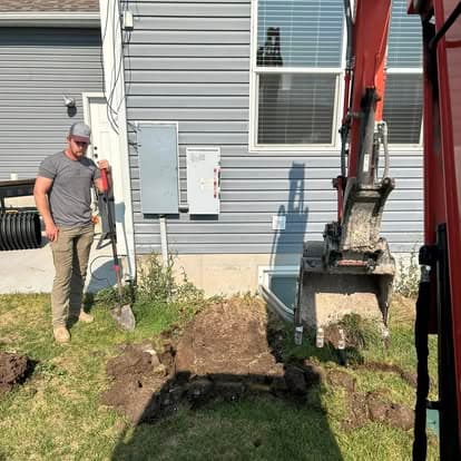 Man supervising an excavation near a house, with a mini excavator and soil mound visible.