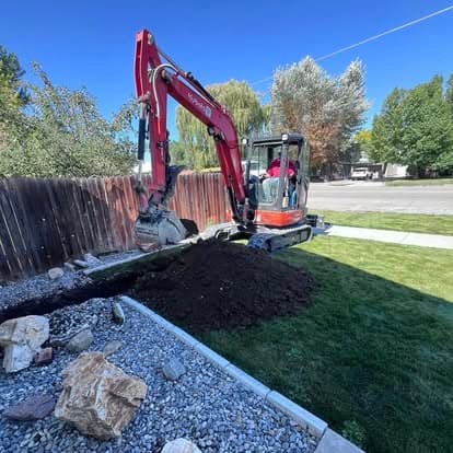 Excavator digging soil in backyard with gravel and rocks under blue sky.